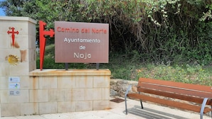 A sign for Camino del Norte Ayuntamiento de Noja, a stone structure with a cross, and a bench in a green, vegetated area.