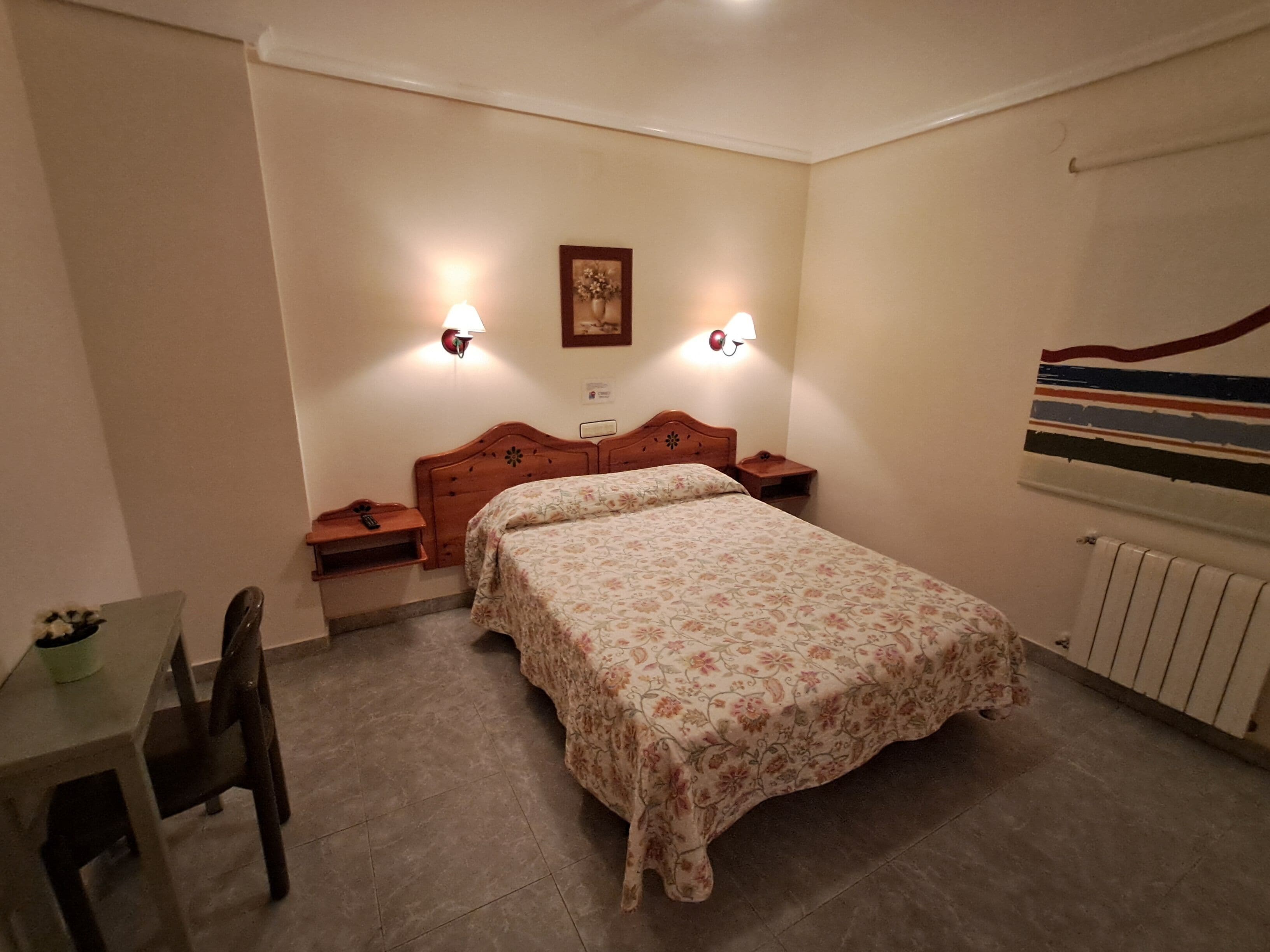 Un dormitorio con cama, mesitas de noche, un sillón, un radiador y dos lámparas de pared.