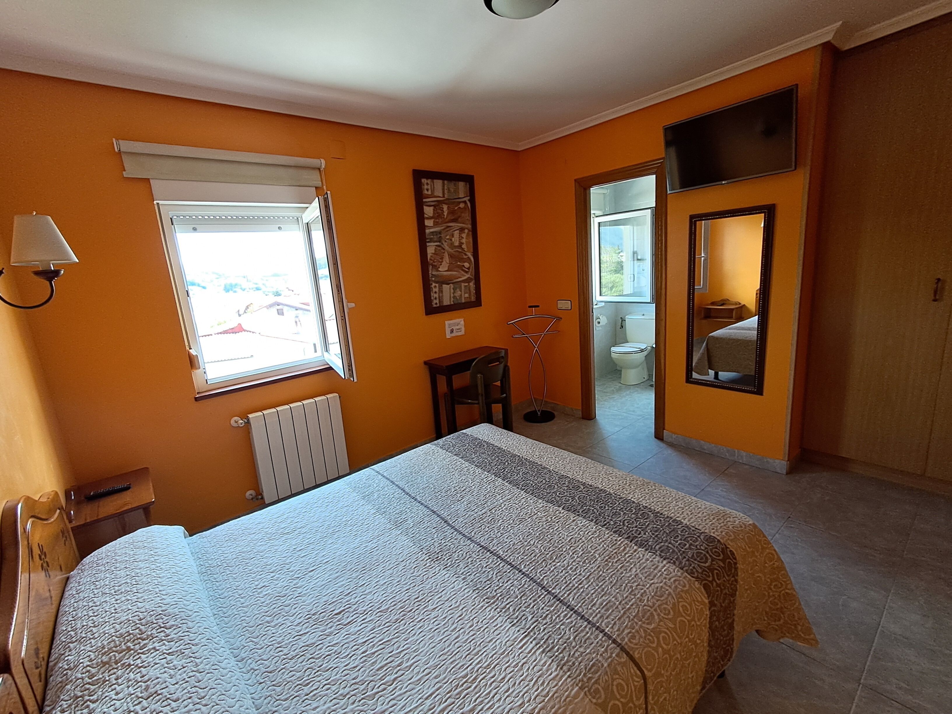 Une chambre à coucher comprenant un lit, un radiateur, une fenêtre, une porte donnant sur une salle de bain et un miroir.