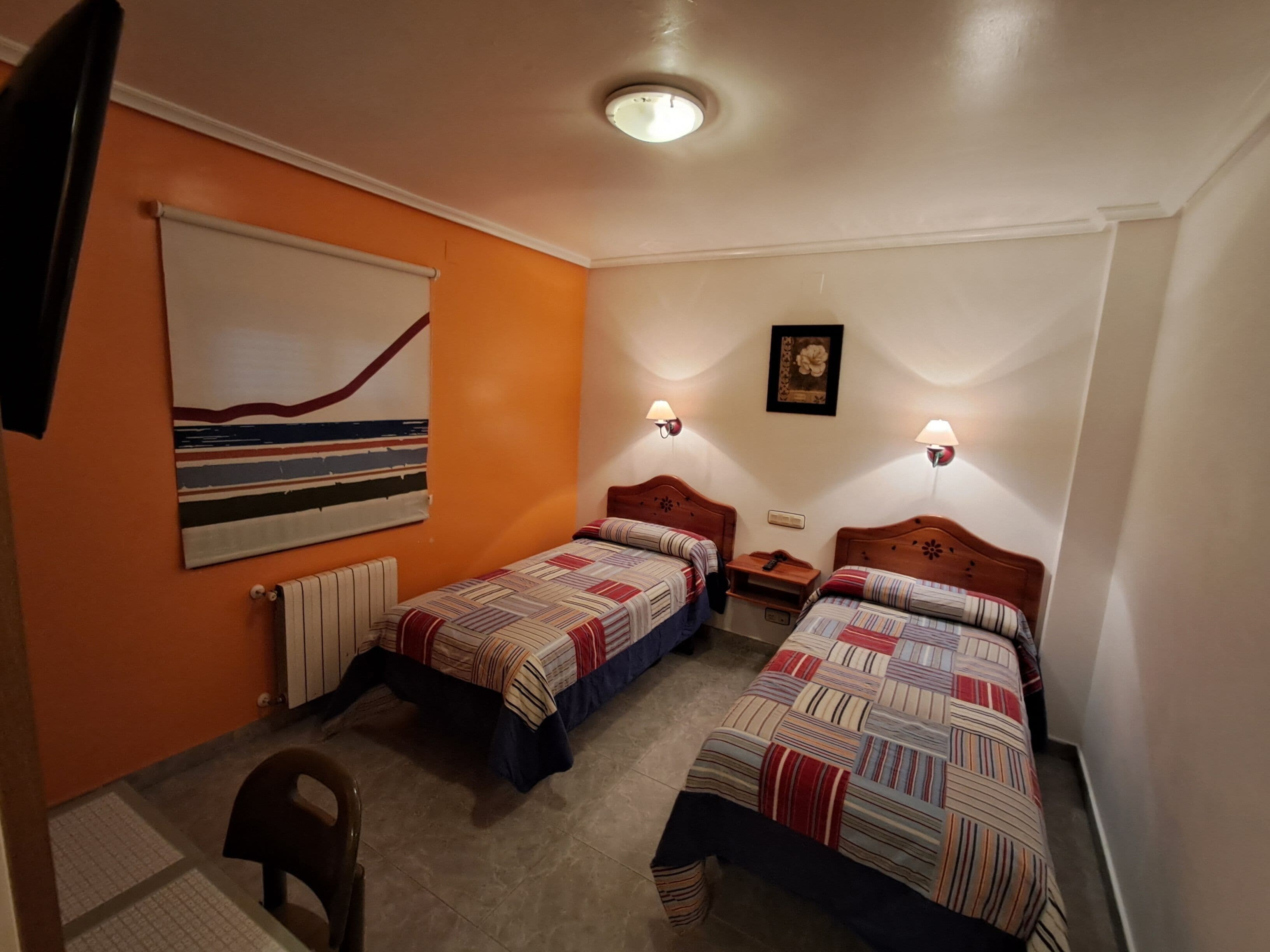 A room with two beds, a radiator, a painting, and two wall-mounted lamps.