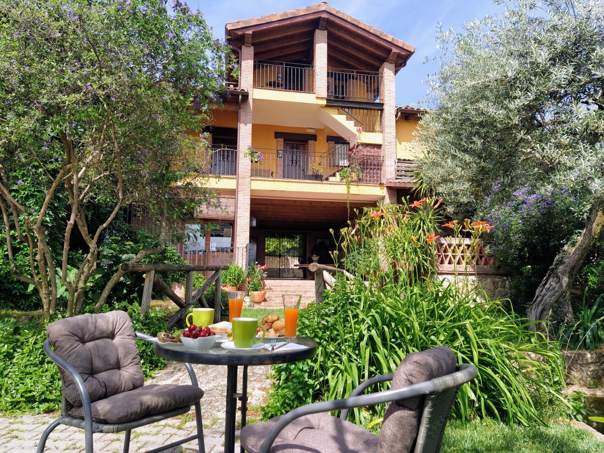 Un bâtiment à deux étages doté de balcons, entouré d’une végétation luxuriante et comprenant un petit espace extérieur aménagé avec des chaises et une table.