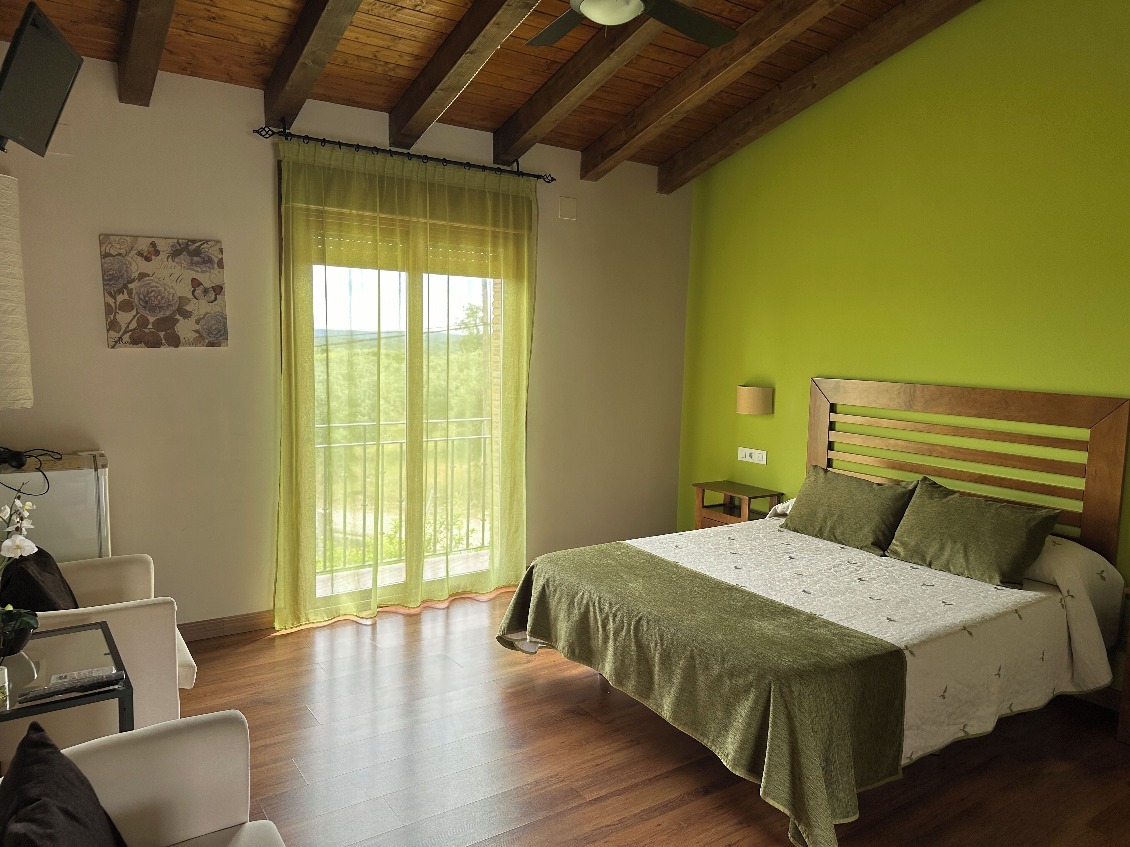 Un dormitorio con techo de madera, una cama con cubrecama verde, una mesita de noche con una lámpara y una ventana con cortinas traslúcidas.