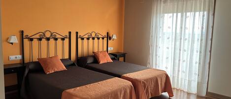A room with two beds, wooden flooring, a window with curtains, and a ceiling with exposed beams.