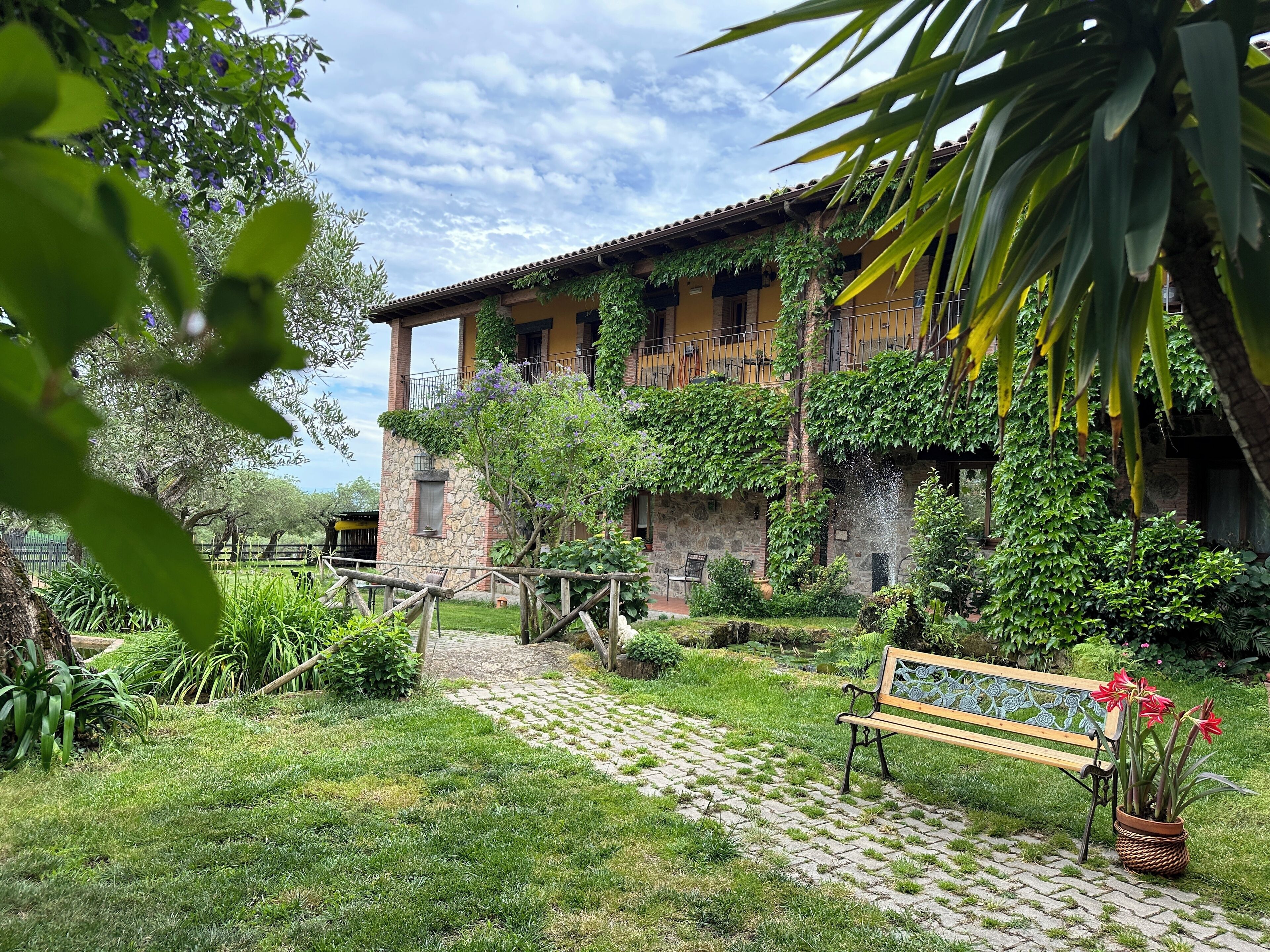 A stone building with a balcony, surrounded by lush greenery and a paved path.