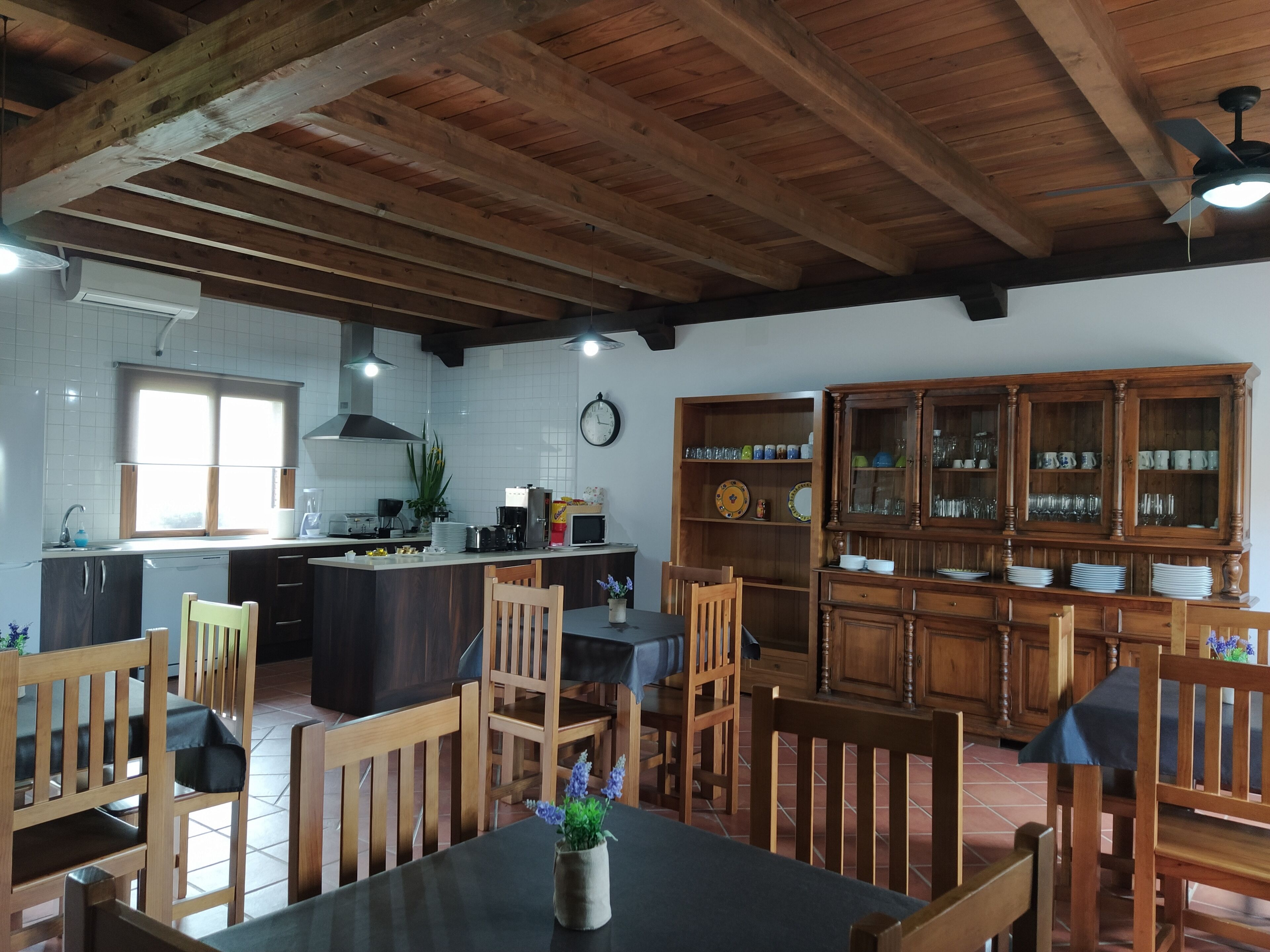 A kitchen with wooden beams, a dining area with wooden chairs, a clock, and a cabinet with dishes.