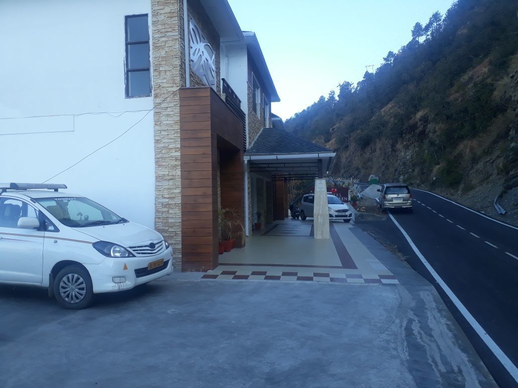 A building with a covered entrance, a white car parked in front, and a road with vehicles.