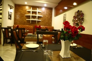Un comedor con mesas dispuestas para dos, un jarrón de rosas rojas y una pared de ladrillo al fondo.