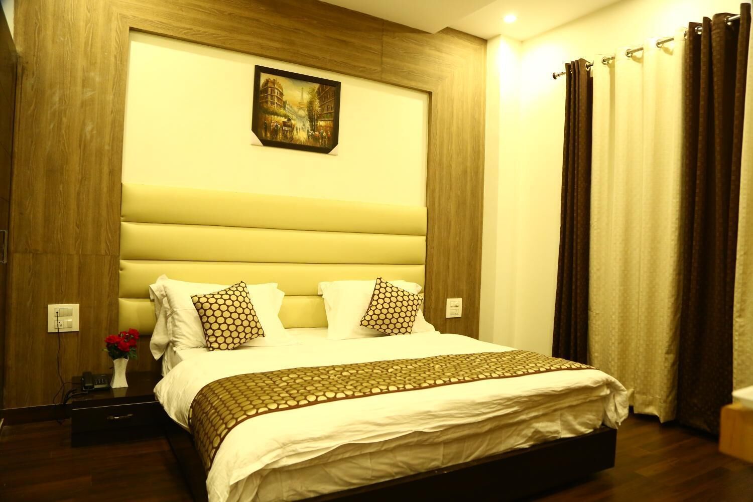 A hotel room with a large bed, a wooden headboard, a painting of a cityscape, and a nightstand with a vase of flowers.