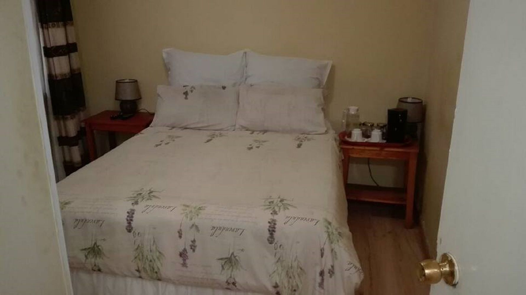 Ein Schlafzimmer mit einem Bett, zwei Kissen, einem Nachttisch mit einer Lampe und einem kleinen Tisch mit Flasche und Tasse.