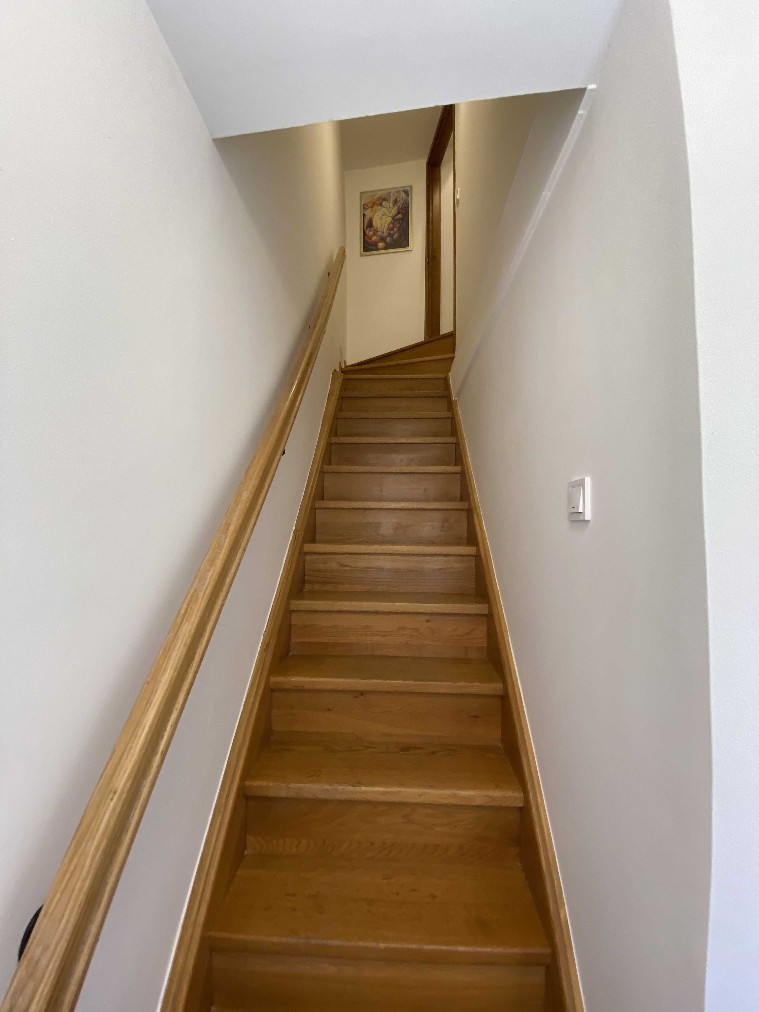 A wooden staircase with a handrail, leading to a landing with a light switch and a framed picture on the wall.