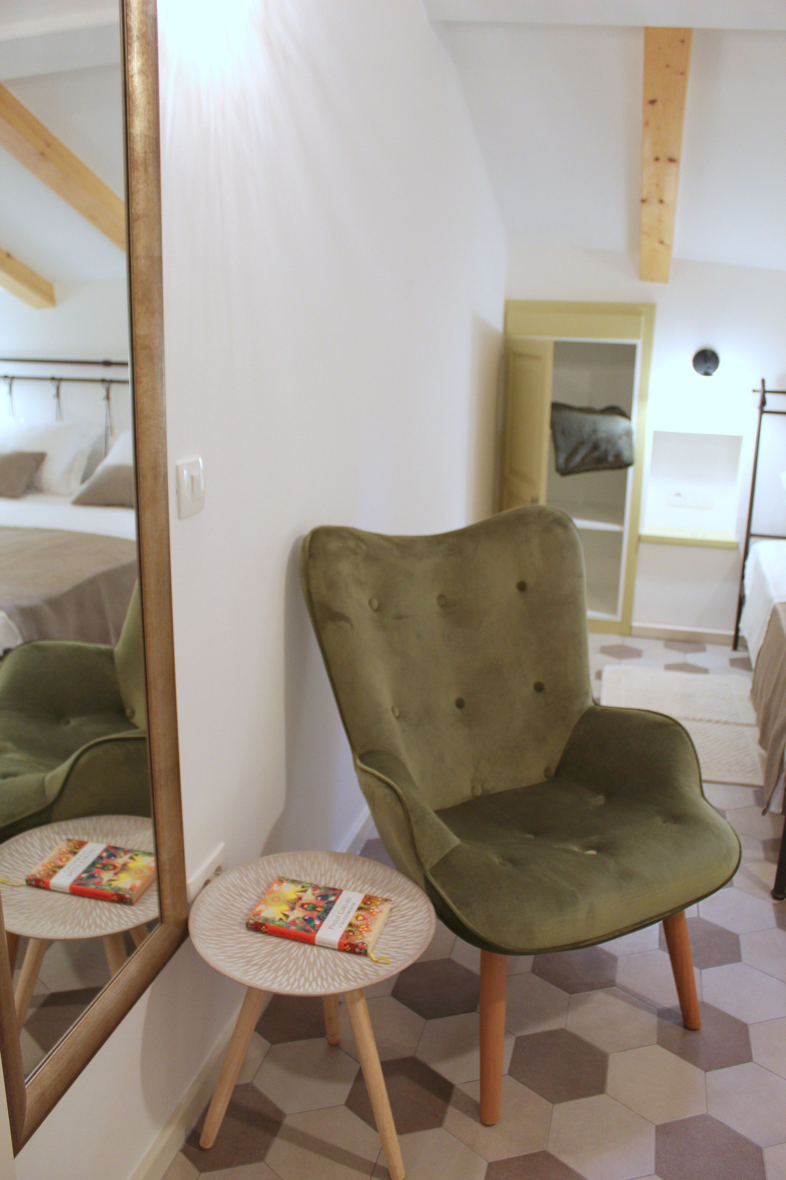 Una habitación con un sillón verde, una mesita redonda con un libro, un espejo grande y una cama al fondo.