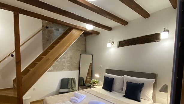 A bedroom with a large bed, wooden beams, a staircase, a small bedside table, and a nightstand. - Noemi's rooms (Rovinj)