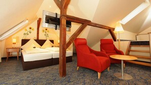 Ein gemütliches Schlafzimmer im Dachgeschoss mit einem Bett, zwei roten Sesseln, einem kleinen Tisch und einer Holzbalkendecke.