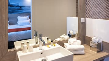 A hotel bathroom with a large sink, mirror, and a bed visible in the background.