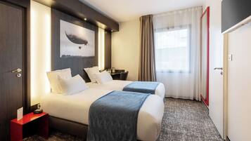 A hotel room with two beds, a nightstand with a clock, a red side table, a wall-mounted artwork, and a large window with curtains.
