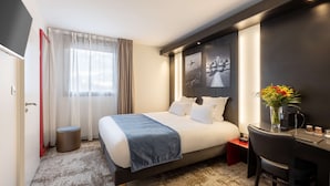 Une chambre d’hôtel moderne avec un grand lit, un bureau et une chaise. On y trouve une fenêtre avec des rideaux, un petit tabouret et un vase de fleurs.