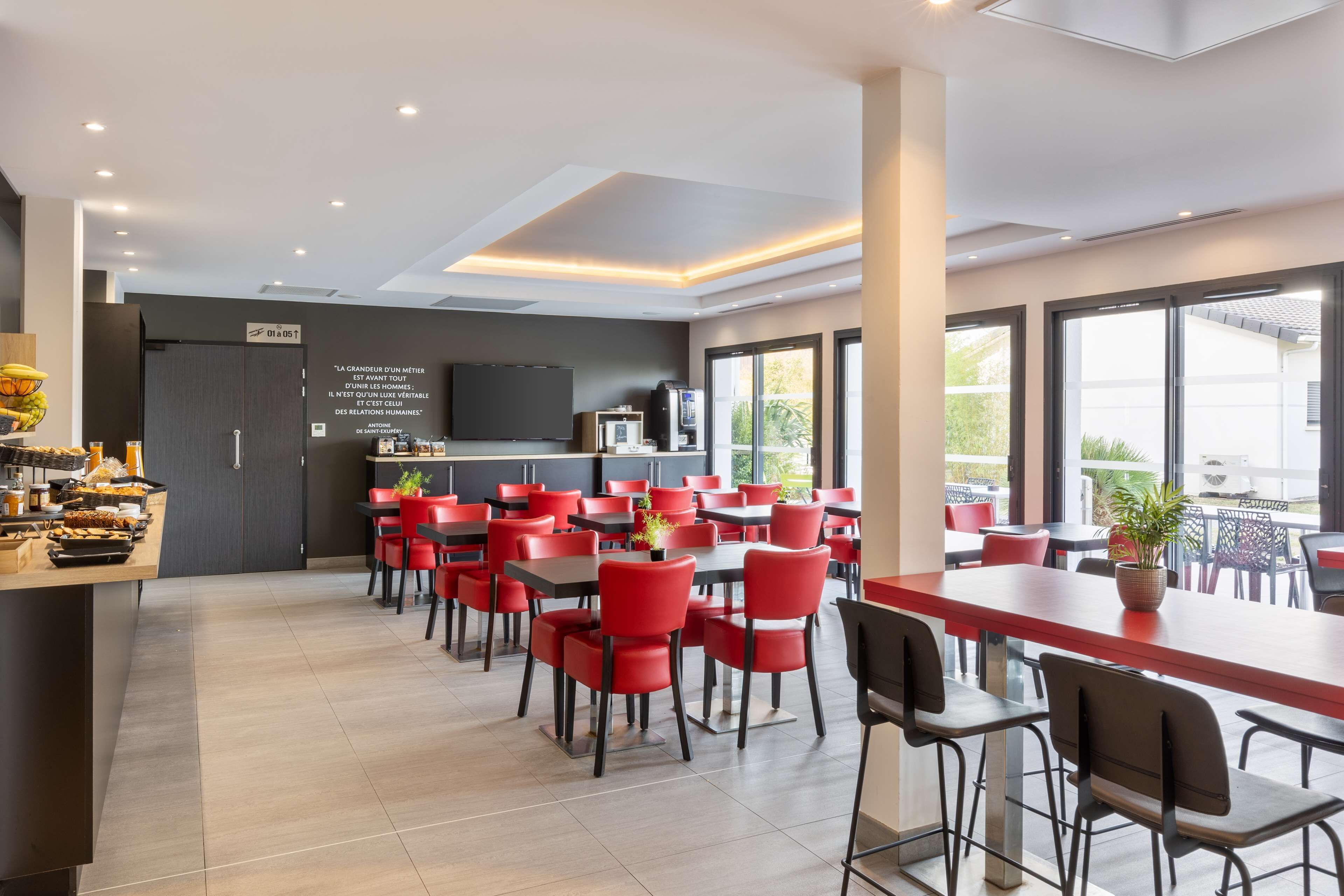 Un espace repas moderne avec des chaises et des tables rouges, un coin cuisine équipé d’un téléviseur et de grandes fenêtres offrant une vue dégagée.