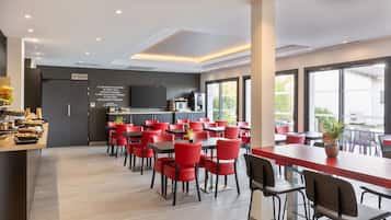 Un espace repas moderne avec des chaises et des tables rouges, un coin cuisine équipé d’un téléviseur et de grandes fenêtres offrant une vue dégagée.
