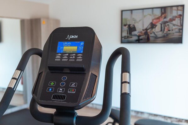 Una bicicleta estática con pantalla digital y panel de control, una cinta de correr y un cuadro en la pared.