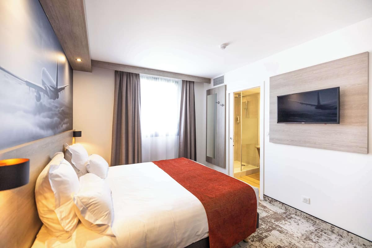 A hotel room with a bed, a TV, a shower area, and a large window with curtains.