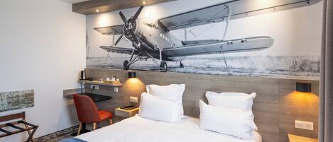 Une chambre d’hôtel avec un lit, un bureau, une chaise et un tableau représentant un avion accroché au mur.