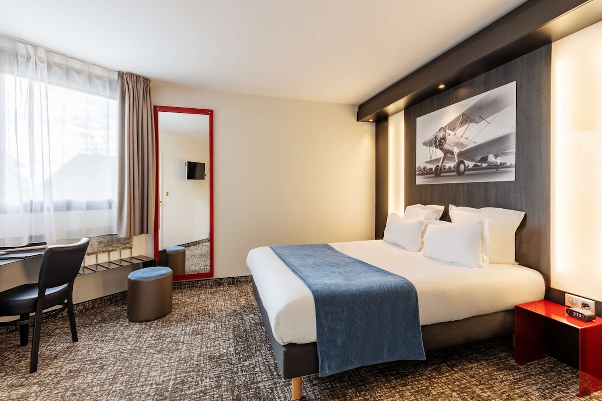 A hotel room with a bed, a desk, a chair, a small stool, and a wall-mounted TV.