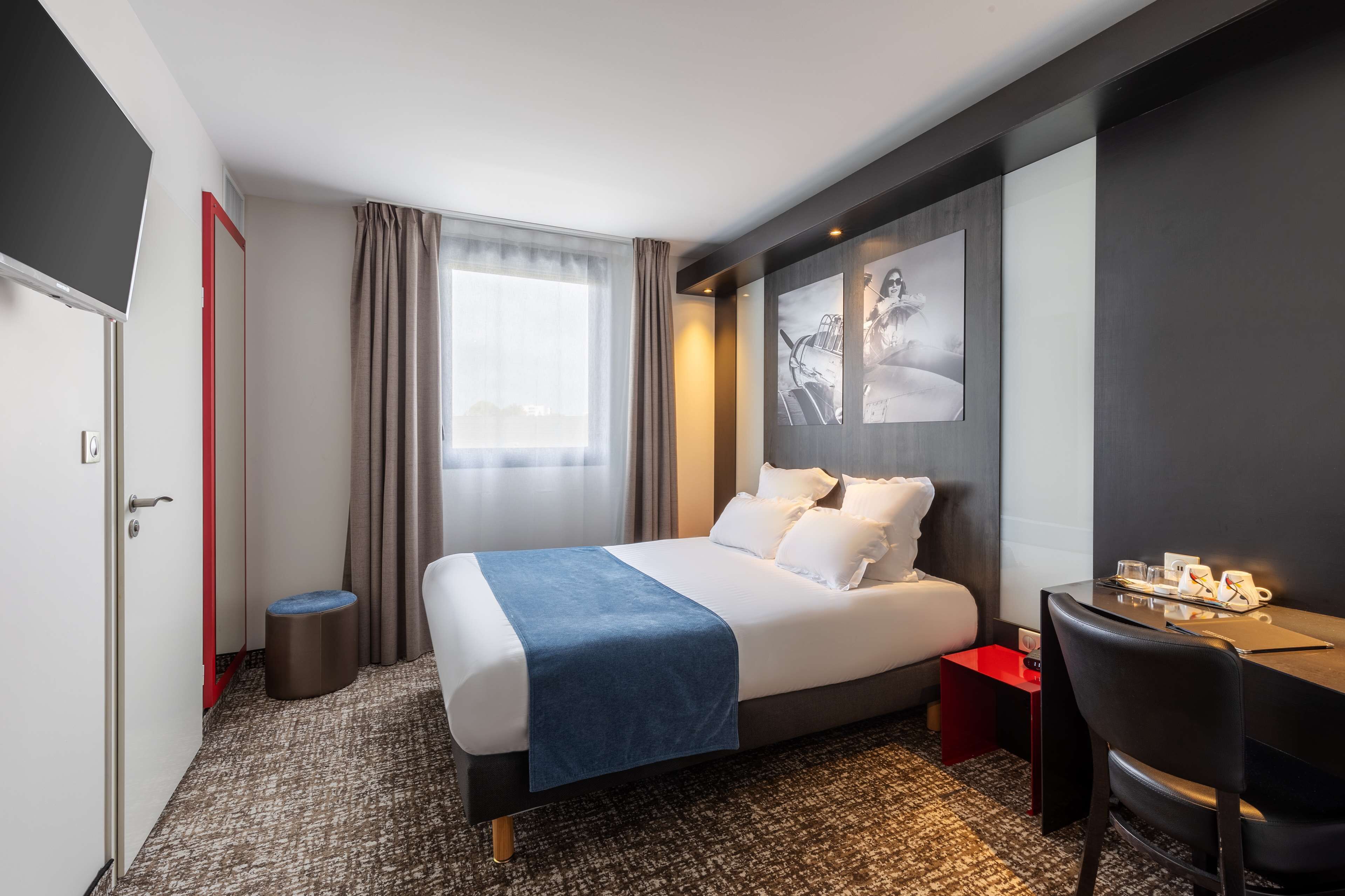 A hotel room with a bed, a desk, a chair, a small stool, a window with curtains, and a wall with framed pictures.