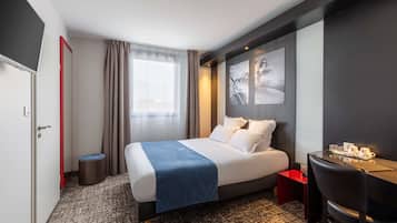 A hotel room with a bed, a desk, a chair, a small stool, a window with curtains, and a wall with framed pictures.