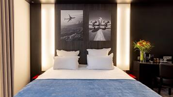 A hotel room with a large bed, two white pillows, a blue throw blanket, a desk with a chair, a vase with flowers, and two framed pictures on the wall.