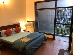 A hotel room with a bed, a nightstand, a lamp, a window with a view of buildings and greenery, and a wooden floor. - Sailors Home (New Delhi)