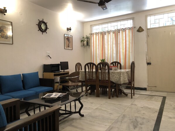 A living room with a blue sofa, a coffee table, a dining table with chairs, a TV, and a ceiling fan. - Sailors Home (New Delhi)