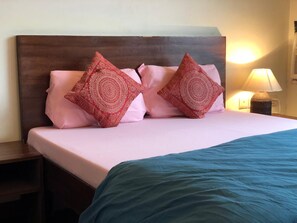 A neatly made bed with pink and red patterned pillows, a teal blanket, and a wooden headboard. - Sailors Home (New Delhi)