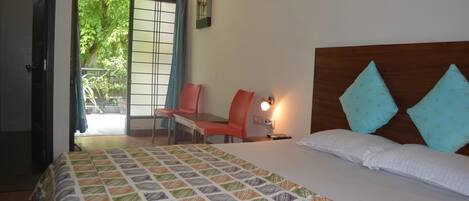 Habitación de hotel con una cama, un escritorio y una silla. Se ve una ventana con vistas a árboles y una puerta que da al exterior.