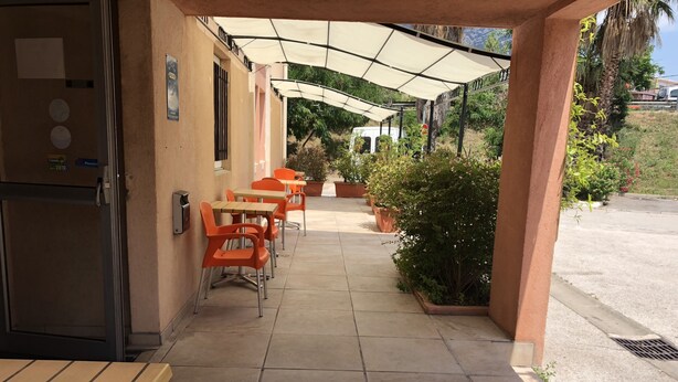 An outdoor patio with orange chairs and tables, a covered area with a white canopy, and a view of a road and greenery in the background. - Hotel Triotel (La Valette-du-Var)
