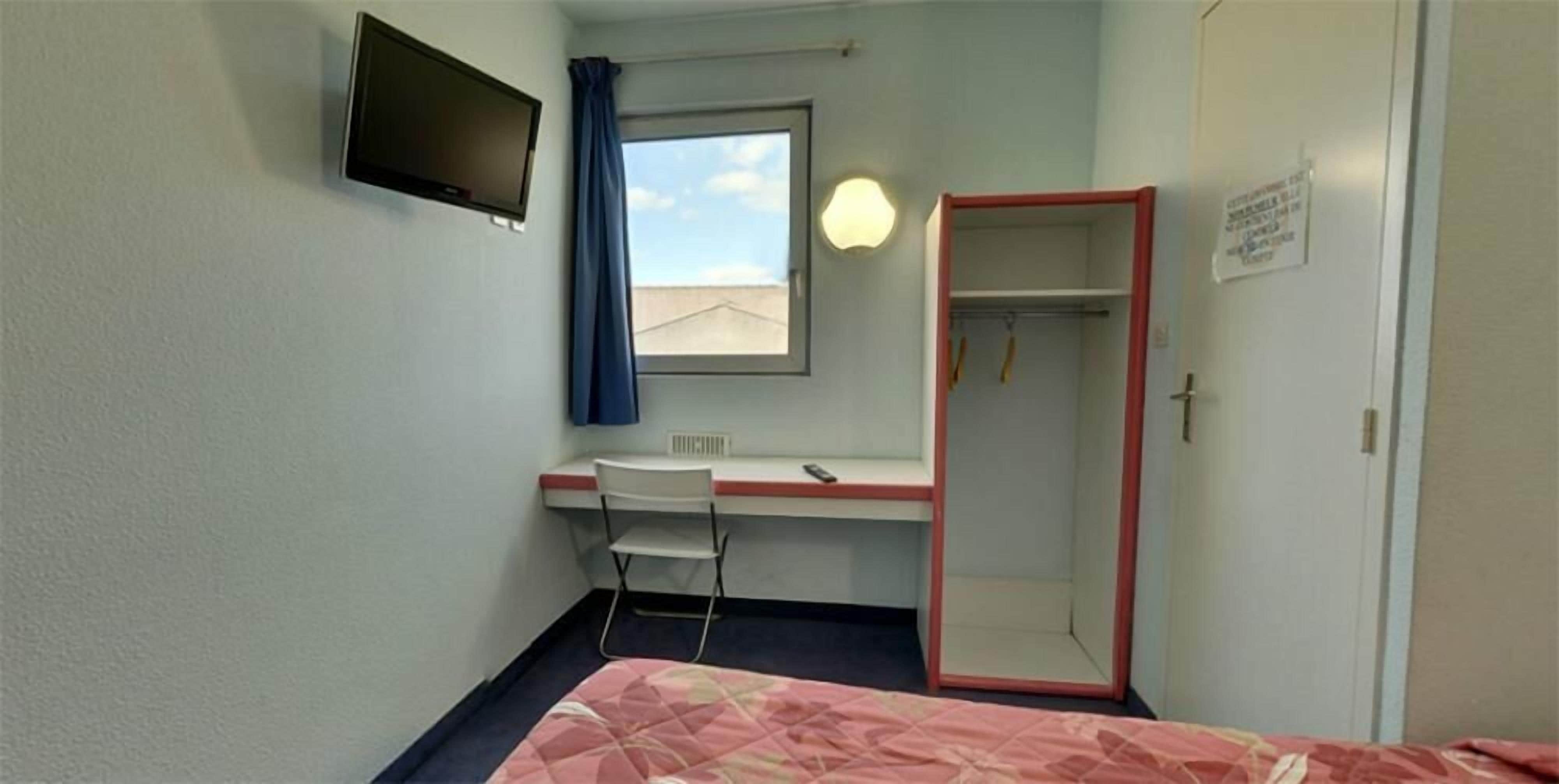 Hotellihuone, jossa on sänky, työpöytä tuolilla, seinälle asennettu televisio, ikkuna verholla ja vaatekaappi.