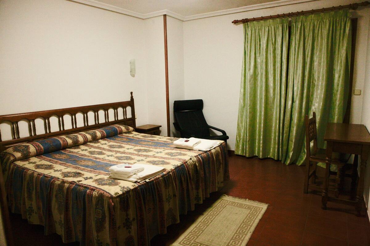 A bedroom with a bed, a chair, a desk, and a green curtain.