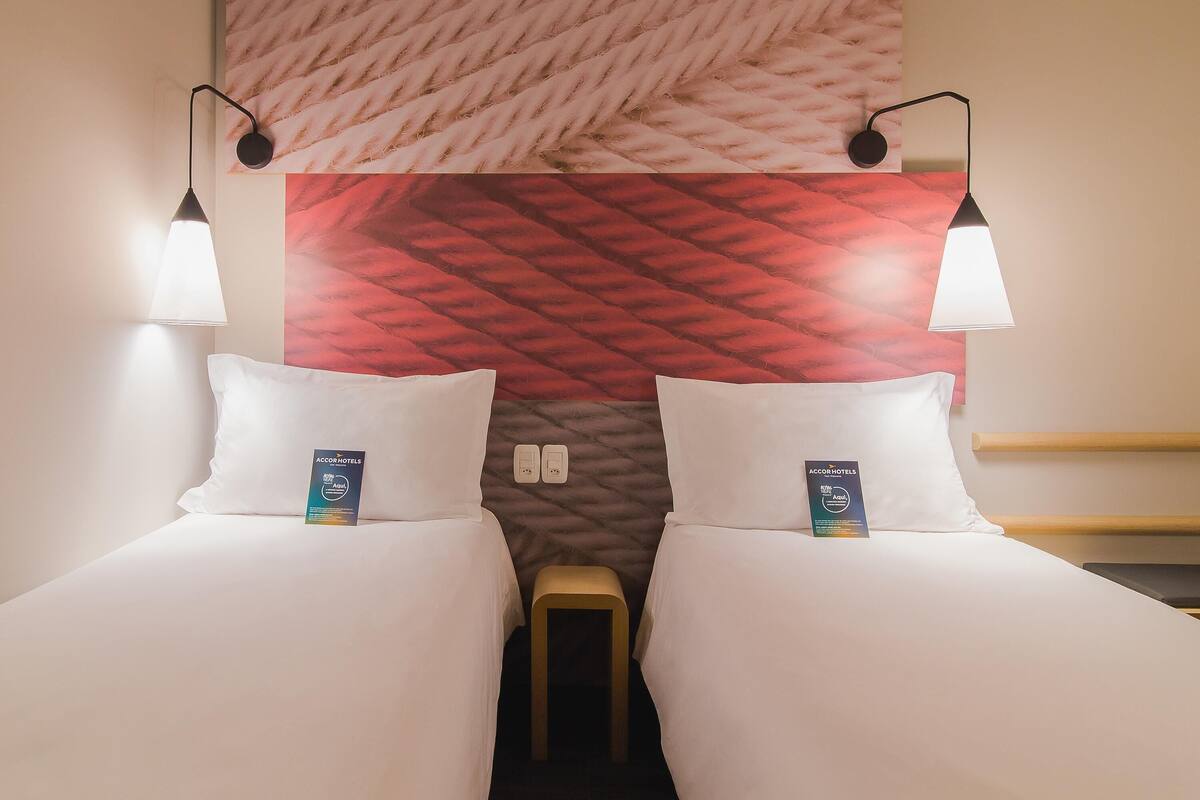 Ein Hotelzimmer mit zwei Betten, einem roten und weißen Muster am Kopfteil, Wandleuchten und einem kleinen Holzschemel.