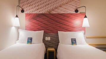 Una habitación de hotel con dos camas, un cabecero con estampado rojo y blanco, lámparas fijadas a la pared y un taburete pequeño de madera.