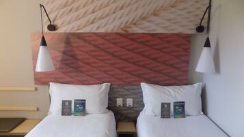 A hotel room with two beds, each with a pillow and a pillowcase, a bedside table, and a wall-mounted lamp.