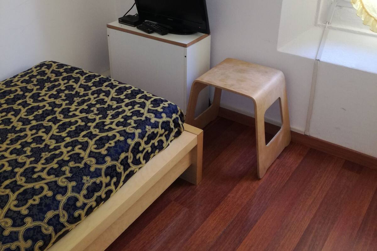 Ein Schlafzimmer mit einem Bett, einem Schreibtisch mit Laptop, einem kleinen Hocker und einem Fenster mit Vorhängen.
