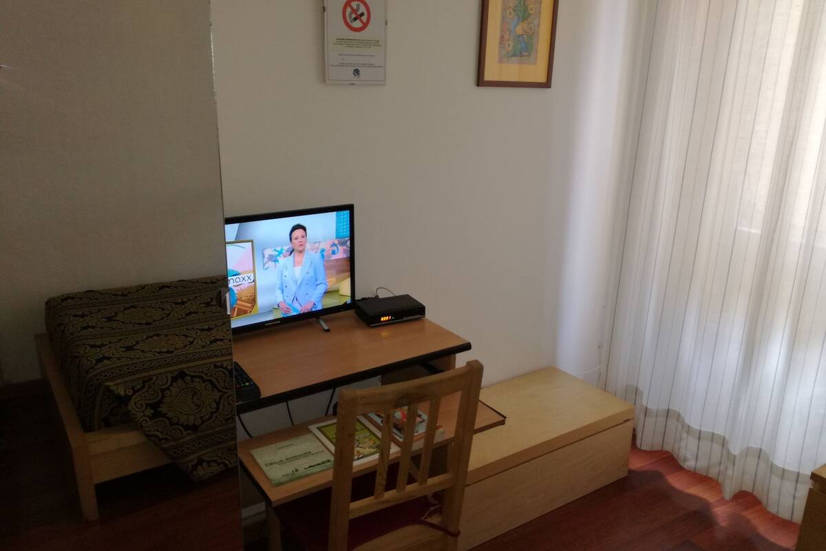 Ein Zimmer mit einem Bett, einem Schreibtisch mit Computer, einem Stuhl, einem gerahmten Bild und einem Fenster mit Vorhängen.