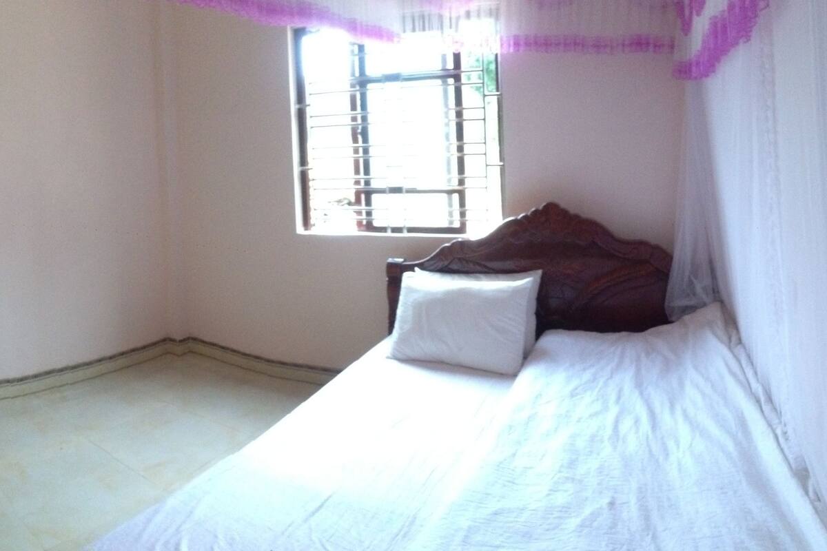 Een kleine, eenvoudig ingerichte kamer met een enkel bed, een raam met tralies en een deur die naar een andere kamer leidt.