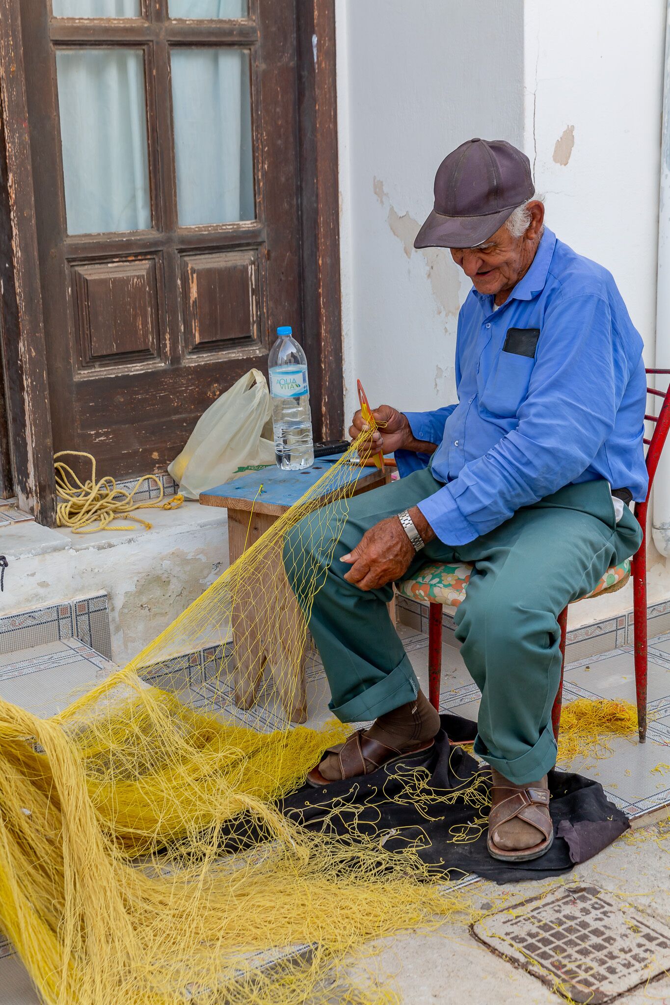 An elderly man sitting on a chair, engaged in a craft activity with yellow threads.