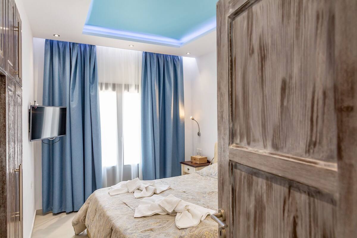 Une chambre à coucher avec un lit, des rideaux, un téléviseur, une table de chevet et une porte en bois.