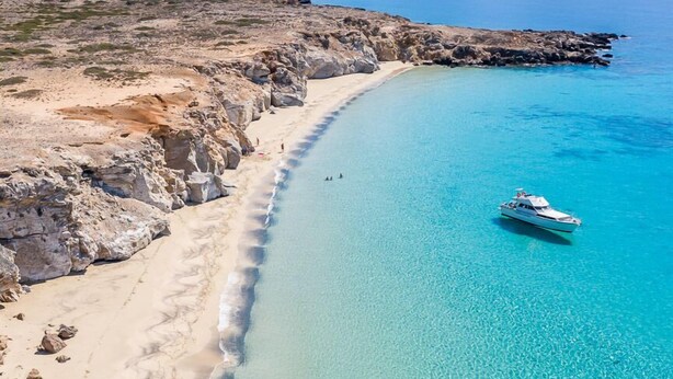 A coastal landscape with a clear beach, rocky outcrops, and a boat on the water. - Theoxenia Suites & Apartments (Kasos)