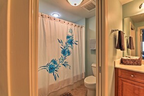 A bathroom with a white shower curtain featuring blue floral designs, a white toilet, a wooden vanity with a basket, and a mirror reflecting the bathroom. - Beachfront Destin Condo w/ Pool & Harbor View! (Destin)