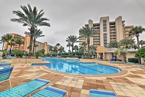 Una piscina con sillas reclinables azules, palmeras y un gran edificio de hotel al fondo.