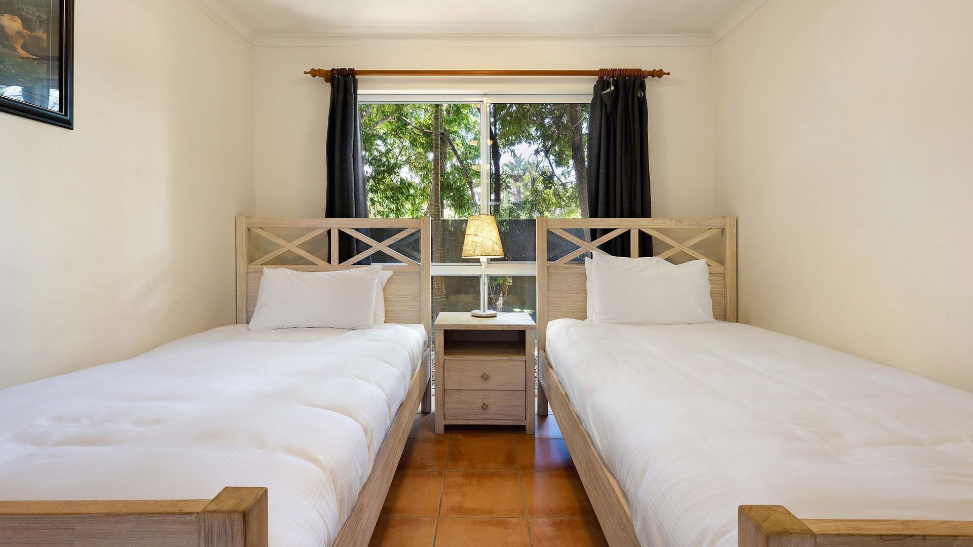 A twin room with two beds, a window with curtains, a nightstand with a lamp, and a view of trees outside.