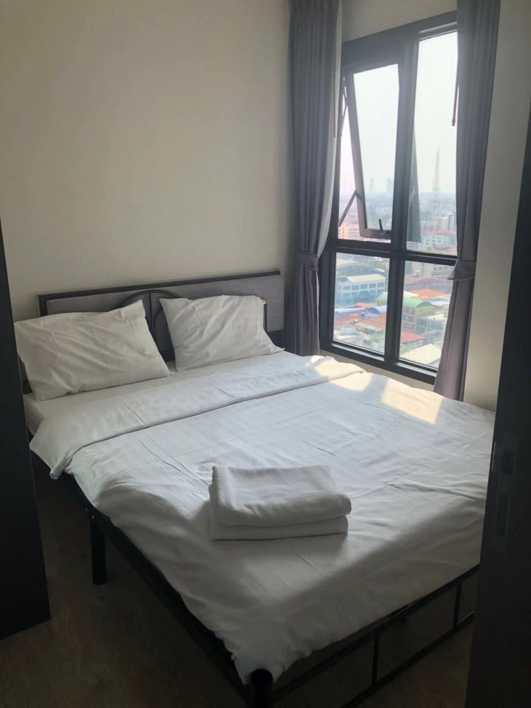 A neatly made bed in a room with a large window, curtains, and a view of buildings outside.