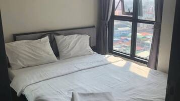 A neatly made bed in a room with a large window, curtains, and a view of buildings outside.